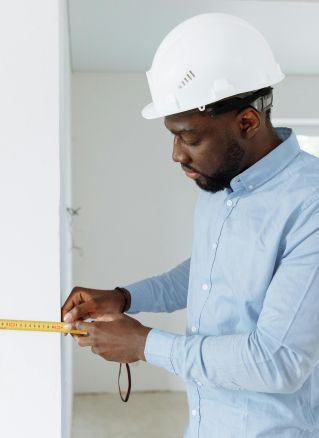 Construction worker measuring a wall indoors, wearing a white hard hat and blue shirt.