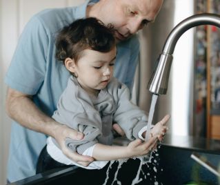 A father tenderly helps his child wash hands in the home kitchen sink.